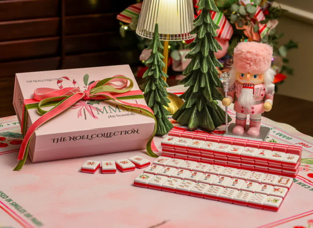 The Noel Collection gift box tied with pink and green ribbon, beside pink-and-red mahjong tiles, a pink nutcracker and ceramic Christmas trees on the Noel mat