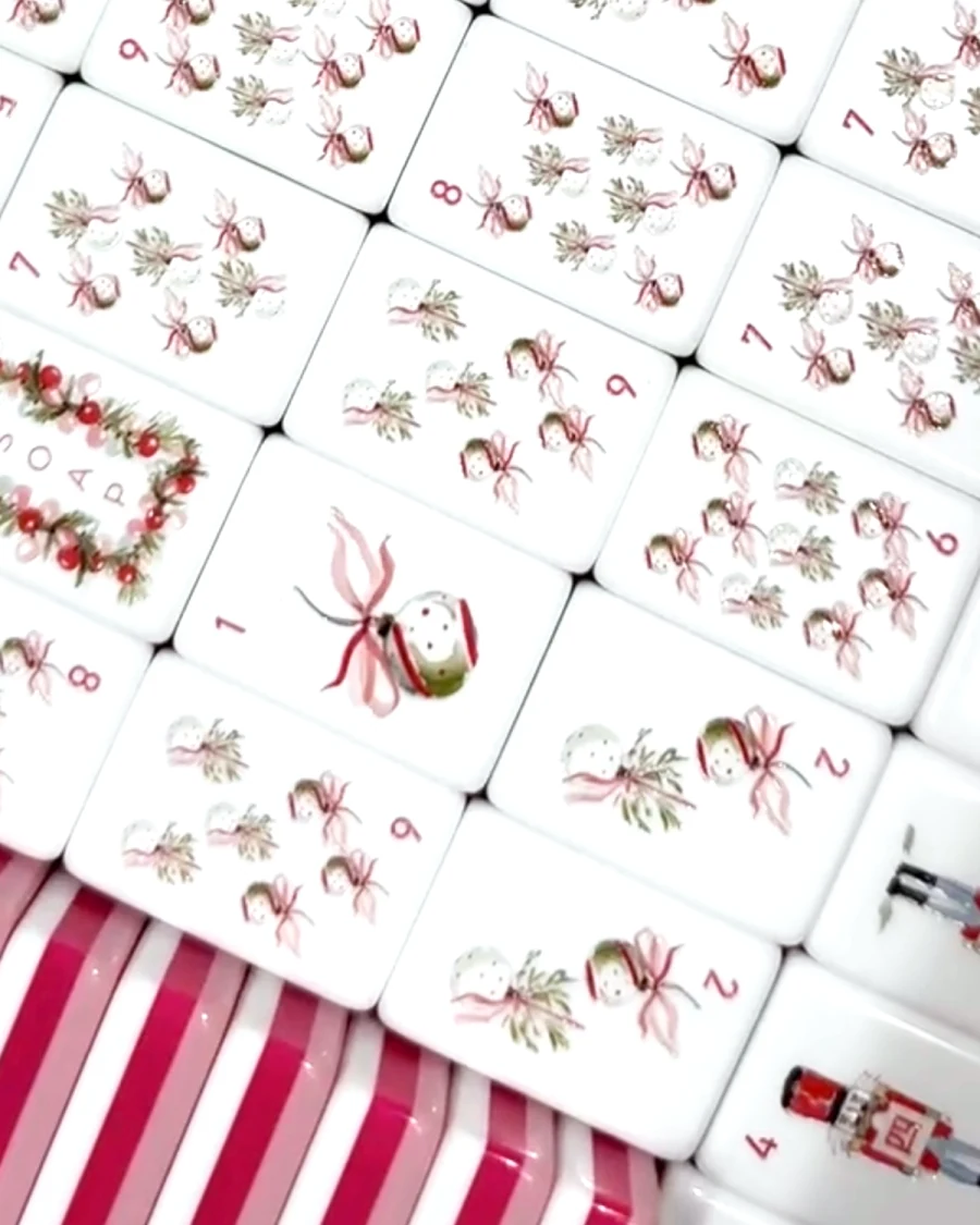 The Noel Collection tiles laid out — numbered suits drawn as ribboned ornaments and sprigs of greenery, with a holly-wreath dragon tile, on a pink striped surface.