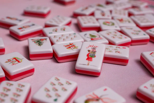 Scattered Noel Collection tiles on a pink cloth, with a joker tile — a woman in a red coat carrying a stack of gifts — in focus among Christmas-tree, dragon and suit tiles.