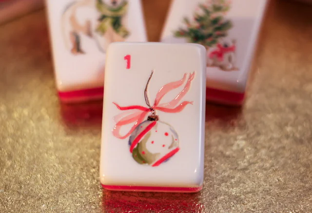 A single Noel Collection “1” tile: a hand-painted Christmas ornament tied with a pink ribbon, on a gold surface with more tiles behind it.